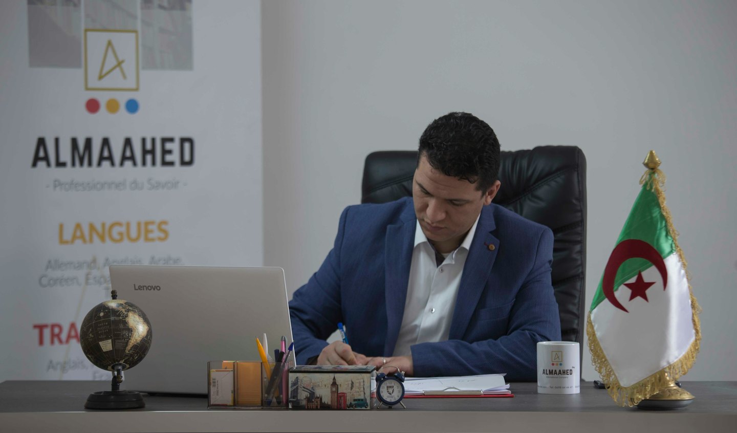 Man in a blue suit writing at his desk.